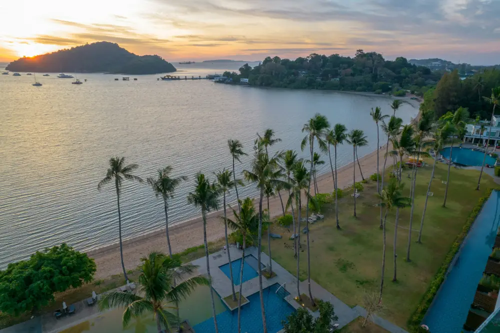Aerial view of beachfront hotel with palm trees, pool, and ocean at sunset