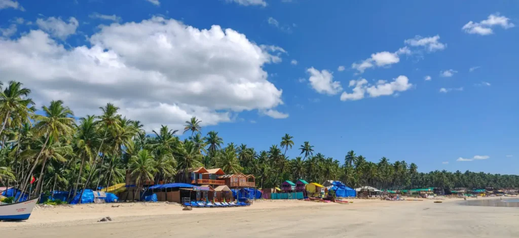 Wide view of Agonda Beach in Goa with palm-lined shoreline, colorful beach huts, soft sand, and a peaceful tropical coastal atmosphere under a bright blue sky
