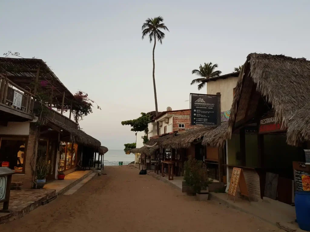 Peaceful coastal village street with traditional thatched-roof buildings, palm trees, and sandy pathways leading to the beach during early evening.