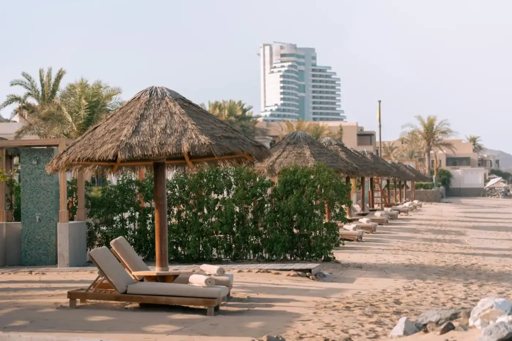 Luxury beach resort cabanas on a sandy shoreline in the UAE with palm trees, sun loungers, and modern seaside hotel buildings in the background.