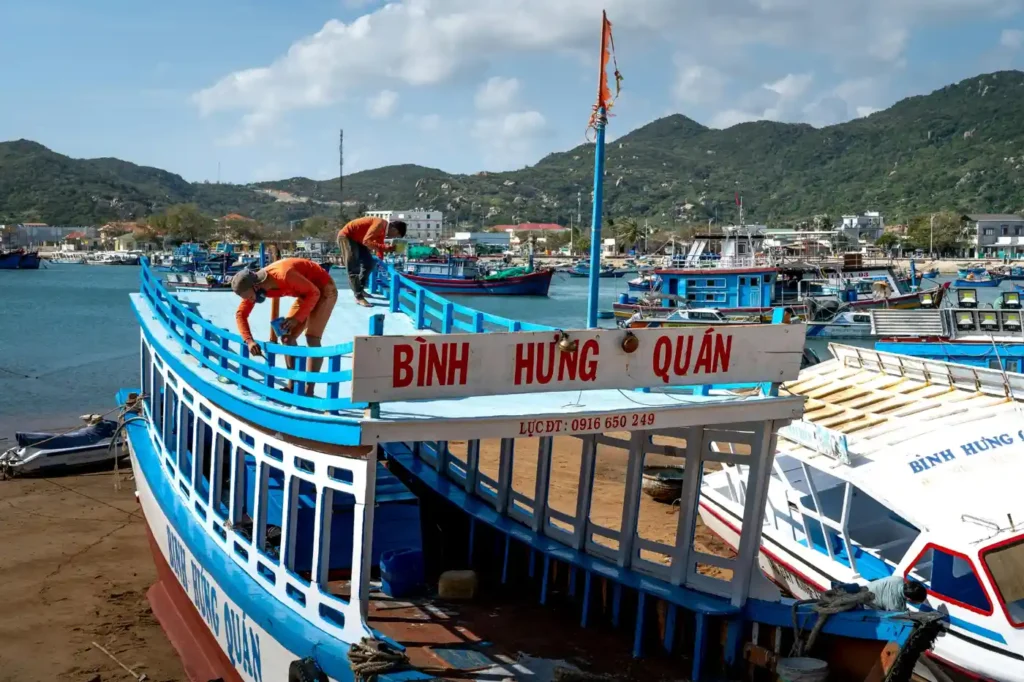 Traditional blue fishing boats docked at Binh Hung Island harbor in Vietnam with green mountains and coastal village in the background.