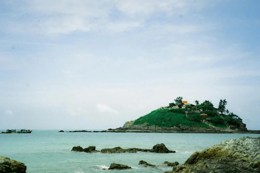 Small green hill island surrounded by calm turquoise water and rocky shoreline at Co To Island in Vietnam.