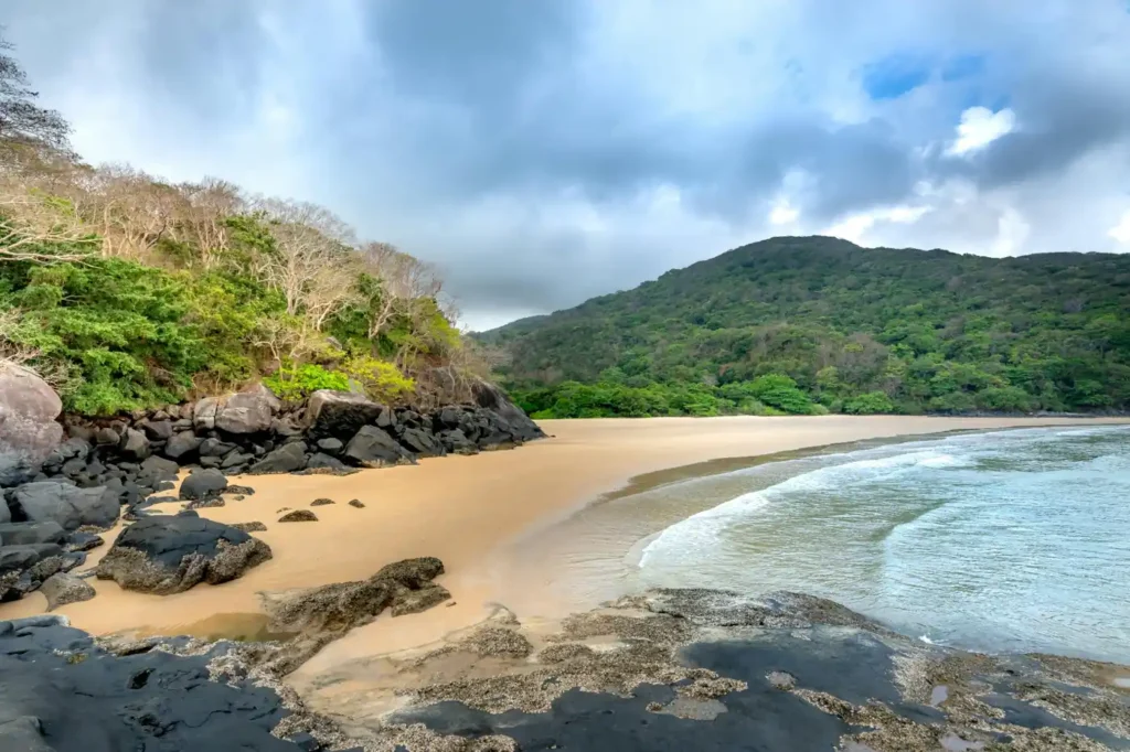 Hidden beach in Con Dao National Park with rocky shoreline, calm waves, forested hills, and cloudy tropical skies in Vietnam