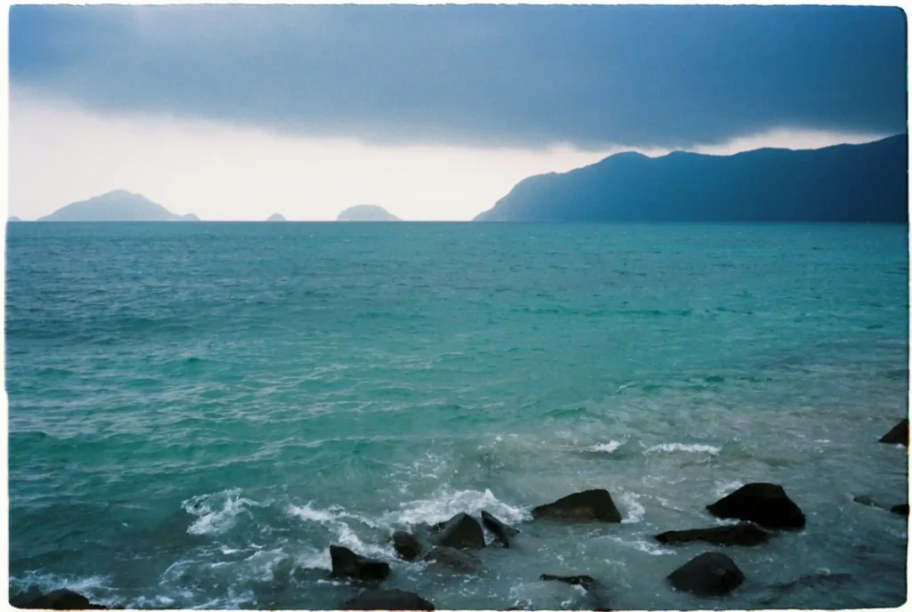 Stormy turquoise sea and rocky shoreline at Con Dao Island in Vietnam with distant island silhouettes under dark clouds.