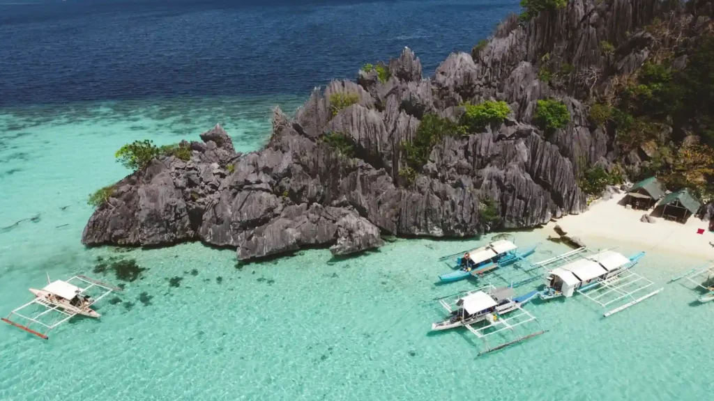 Aerial view of El Nido limestone cliffs surrounded by crystal-clear turquoise water and traditional Filipino boats near a tropical beach in Palawan.