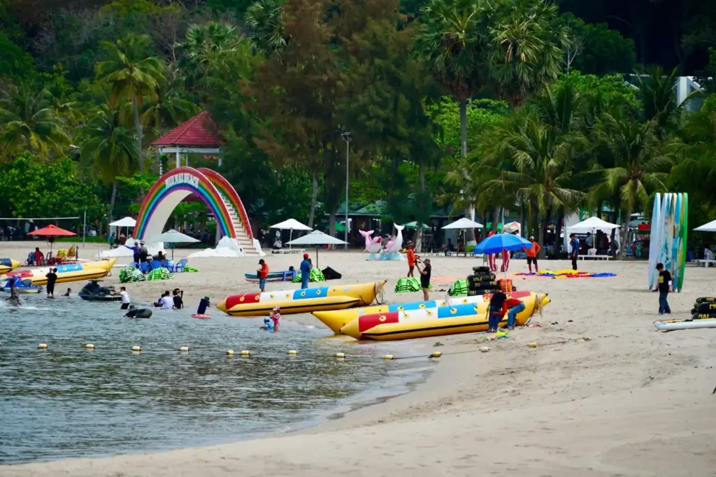 Families enjoying water activities and banana boat rides at a lively beach with palm trees, umbrellas, and seaside attractions near Corniche Beach Abu Dhabi.