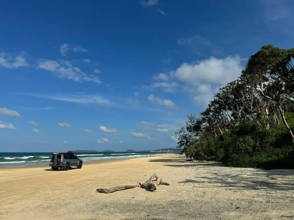 Remote german island beach beach with a parked off-road vehicle, wide sandy shoreline, ocean waves, and coastal trees under a bright blue sky.
