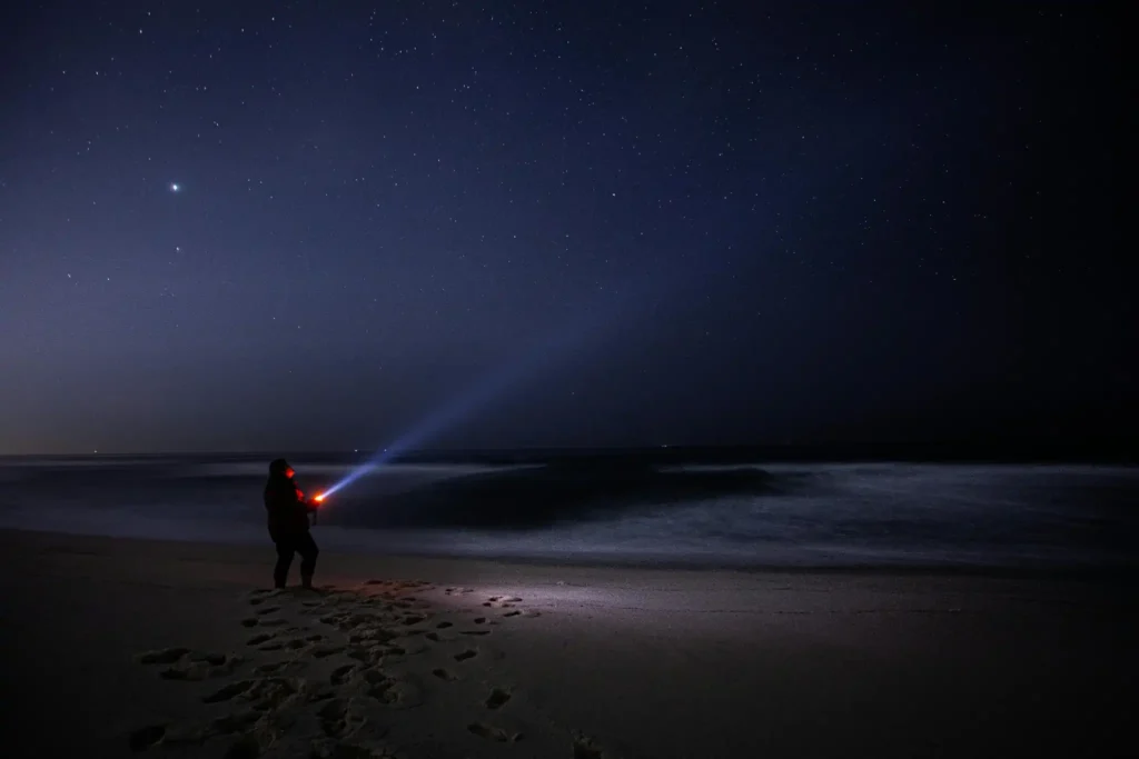 Person standing on a beach at night shining a flashlight toward the ocean under a starry sky at Hiriketiya Beach