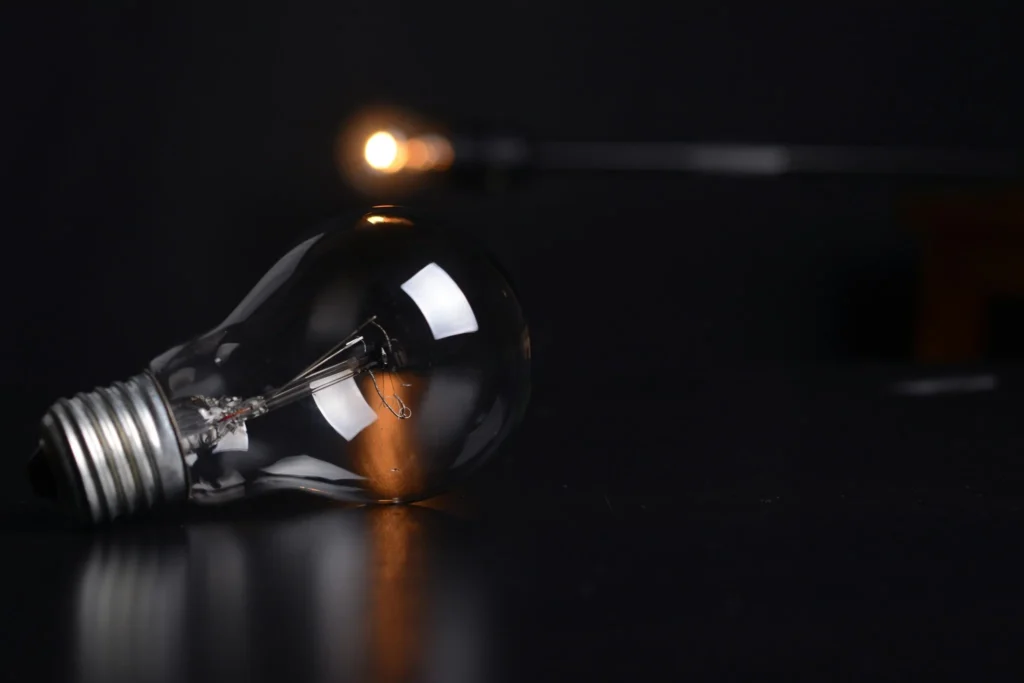 Close-up of a clear incandescent light bulb lying on a dark reflective surface with a blurred glowing light in the background.
