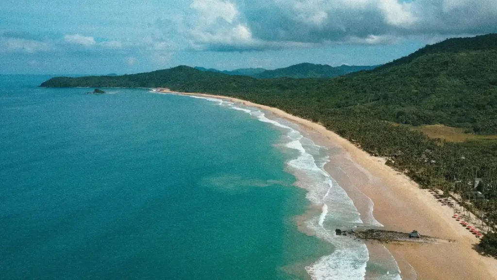 Aerial view of Long Beach near Port Barton with turquoise ocean water, an endless sandy shoreline, tropical forest, and rolling green hills along the coast.
