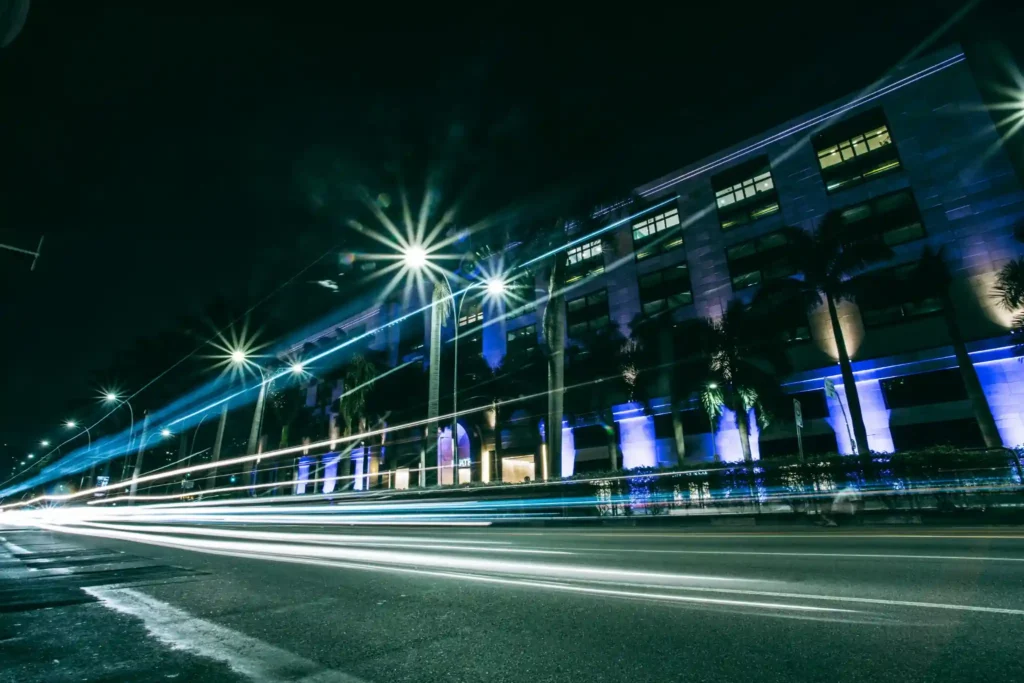 Night view of a modern beachfront city street with long-exposure light trails, illuminated buildings, and palm trees along the road.