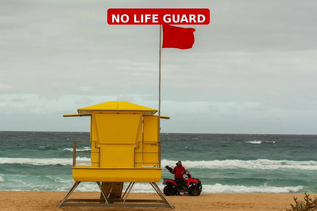 Yellow lifeguard tower with a red warning flag on a rough beach shoreline under cloudy skies, indicating no lifeguard supervision.