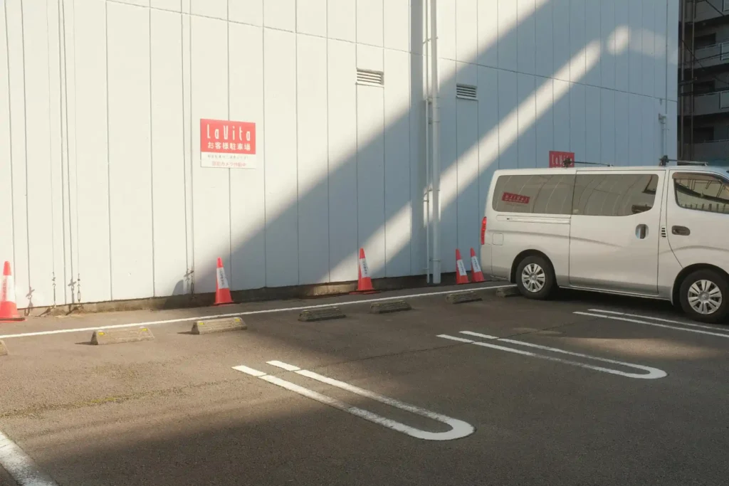 Small urban parking area with a white van, traffic cones, and long afternoon shadows beside a commercial building in Japan.