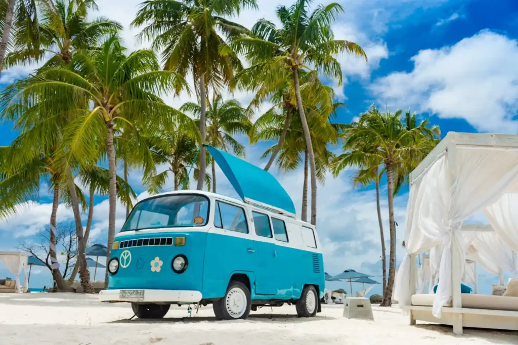 Blue vintage camper van parked on a tropical beach surrounded by palm trees and white sand under a bright sky.