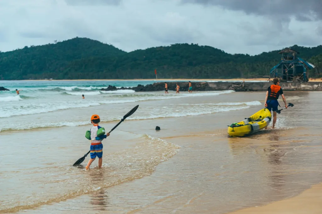 Travelers carrying kayaks along the shoreline of a tropical beach in Port Barton with mountains and cloudy skies in the background.