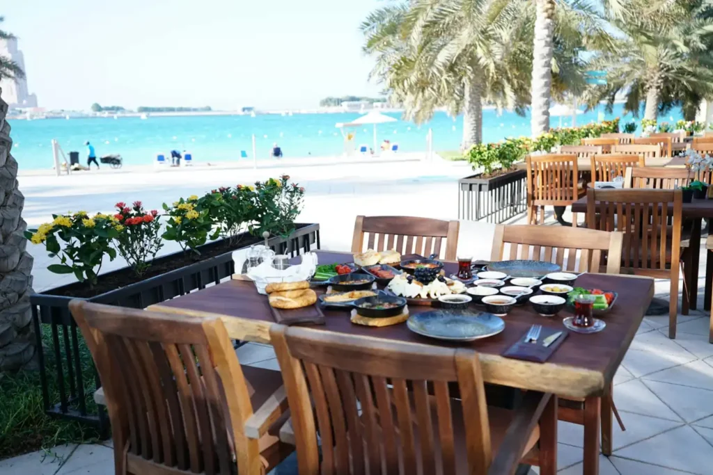 Outdoor beachfront restaurant near Corniche Beach Abu Dhabi with wooden dining tables, traditional breakfast dishes, palm trees, and turquoise sea views in the background.