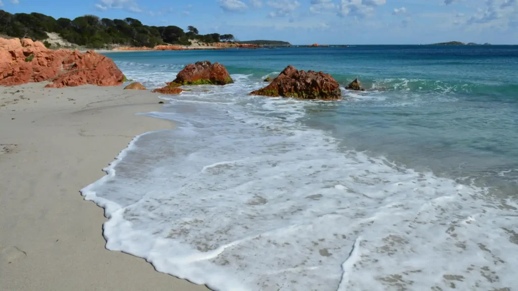 Scenic tropical beach with turquoise water, soft white sand, gentle ocean waves, and reddish coastal rocks under a bright blue sky