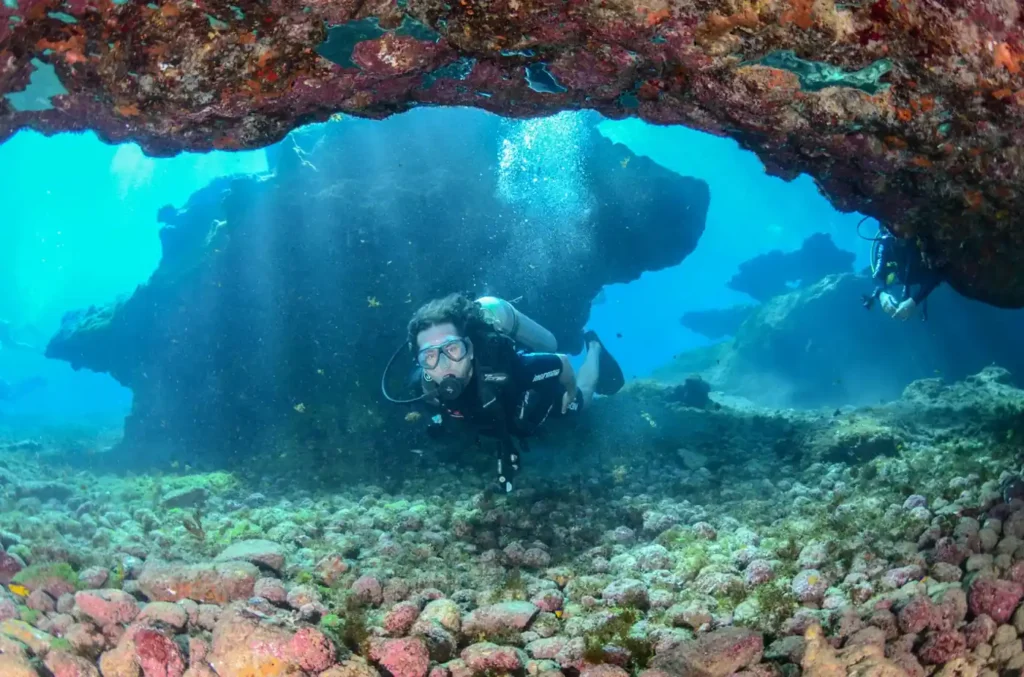 Scuba diver exploring an underwater cave and coral-covered rock formations in the clear blue waters around Koh Kood, Thailand.