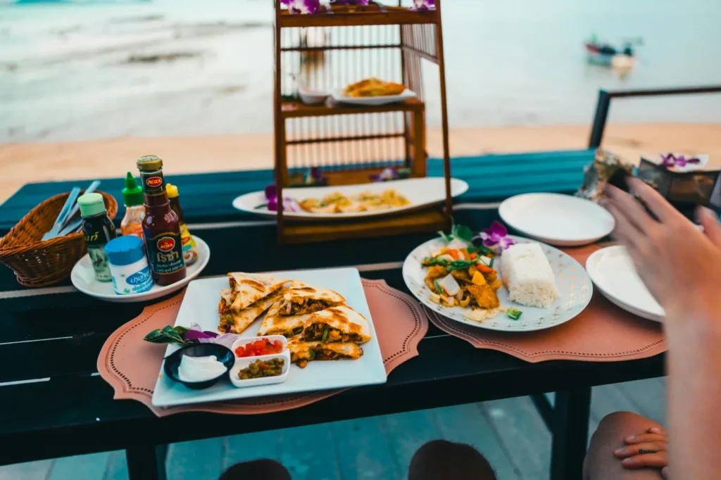 Beachfront dining setup with Thai food, rice dishes, sauces, and tropical decor overlooking the ocean at a seaside restaurant in Thailand.