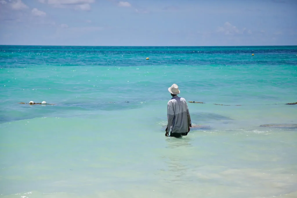 Person standing in shallow turquoise ocean water at a peaceful tropical beach under a bright blue sky.