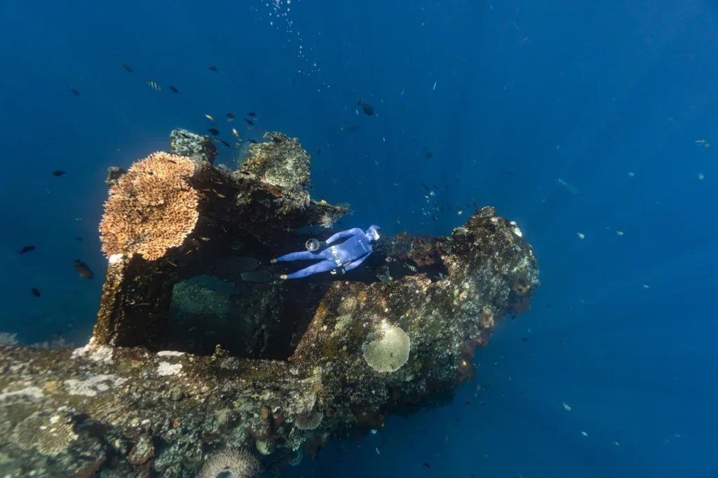 Scuba diver exploring a colorful coral reef and underwater shipwreck in the clear blue waters of the UAE.
