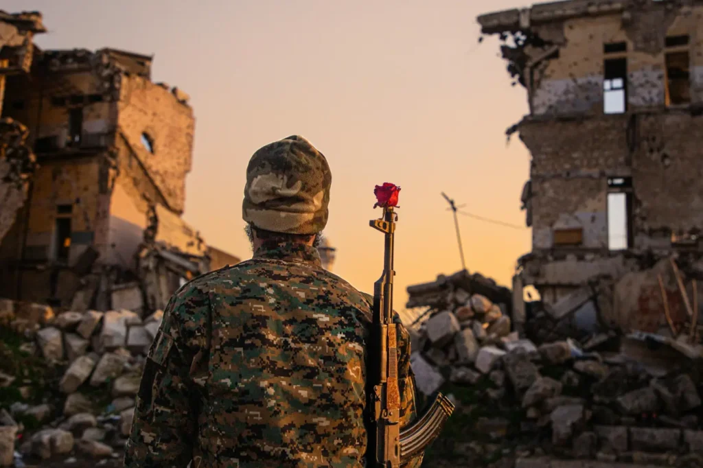 Armed soldier standing among destroyed buildings at sunset with a red flower attached to a rifle, symbolizing conflict, resilience, and the human impact of war.