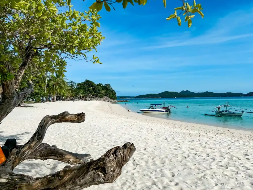 Peaceful white sand beach in Port Barton, Palawan with turquoise water, tropical trees, and traditional boats anchored near the shoreline.