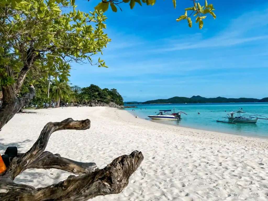 White sandy tropical beach with turquoise water, anchored boats, lush palm trees, and clear blue skies at a peaceful island destination.