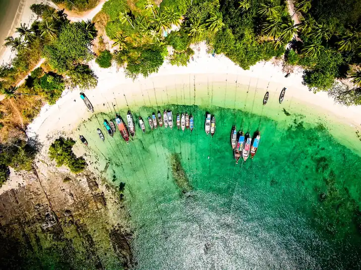 Aerial view of longtail boats anchored in clear turquoise water beside a white sandy beach lined with palm trees. 