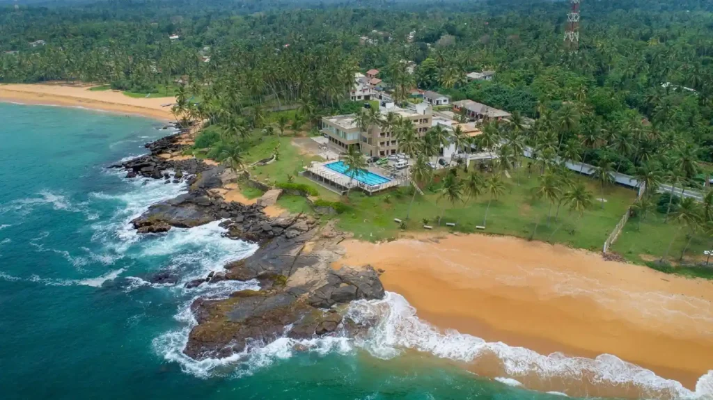 Aerial view of a tropical Sri Lankan beach resort with turquoise water, golden sand, rocky shoreline, palm trees, and nearby buildings.