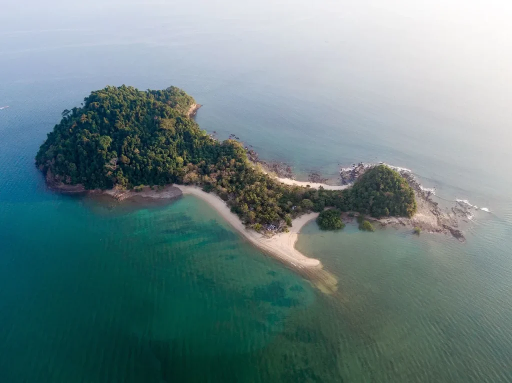 Aerial view of a small tropical island with forested hills, curved sandy beaches, rocky shores, and clear turquoise water.