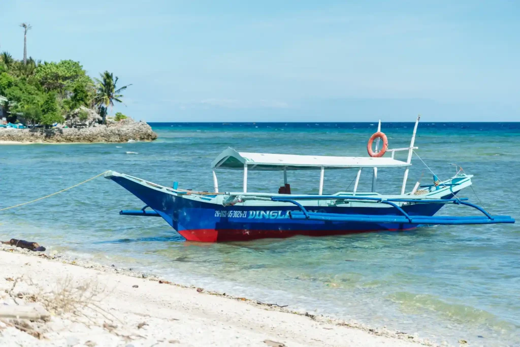 Traditional outrigger boat anchored near a white sandy beach with clear turquoise water and tropical shoreline.