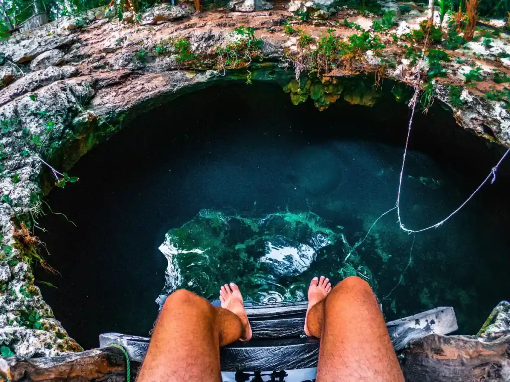 Traveller sitting at the edge of a dark turquoise cave pool with rocky limestone edges, showing a natural swimming hole experience near Anda Beach. 