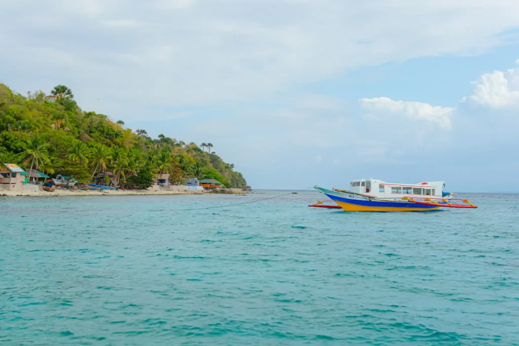Traditional Filipino outrigger boat anchored in clear turquoise water near a tropical island shoreline with palm trees and cloudy skies.