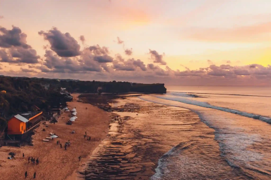 Aerial view of Balangan Beach at sunset with rocky shoreline, gentle waves, and golden reflections across the ocean.