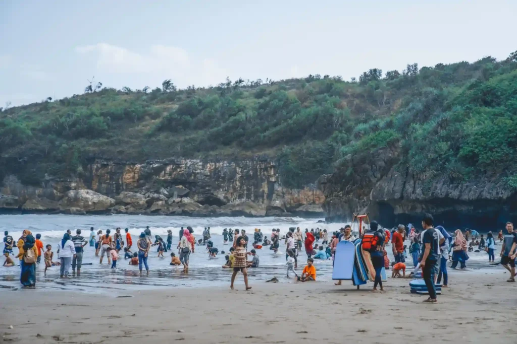 Visitors enjoying Balangan Beach in Bali during a busy day, with families playing in the waves and limestone cliffs in the background.