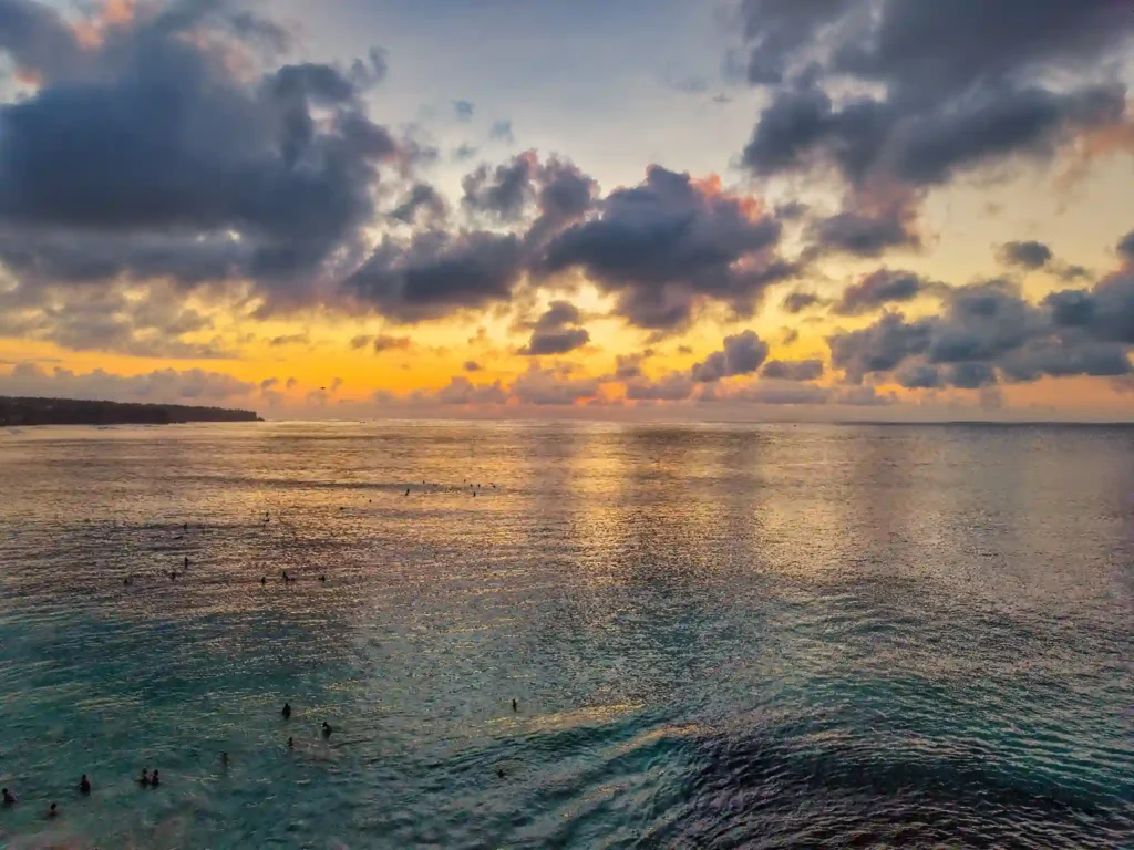 Sunset over the Indian Ocean at Balangan Beach Bali, with dramatic clouds, golden light reflecting on the water, and surfers waiting offshore.
