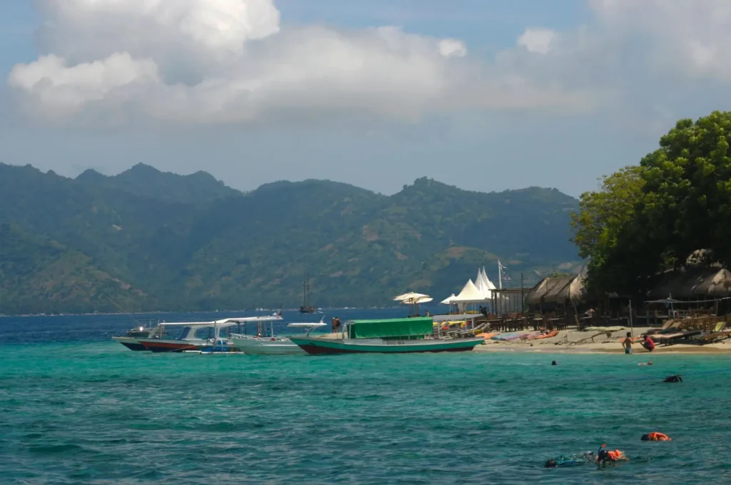 Boats anchored near a tropical beach with snorkelers in clear turquoise water and green mountains in the background.