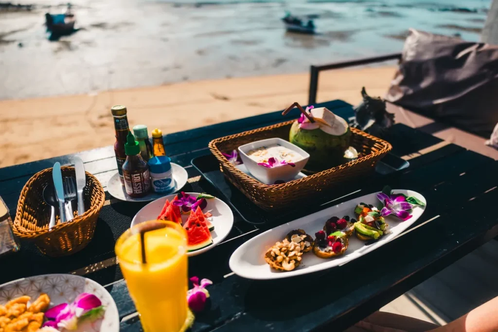 Beachfront breakfast table with tropical fruit, drinks, coconut, and plated snacks overlooking the sea with boats in the background.