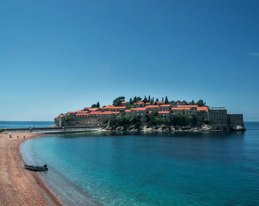 Clear turquoise beach water with a small island village, red-roof buildings, sandy shore, and calm coastal scenery.