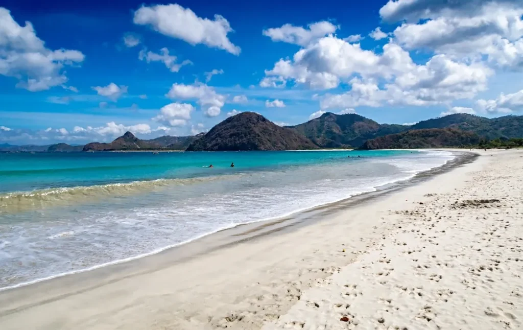 White sandy beach with turquoise water, gentle waves, and green mountains under a bright cloudy sky.
