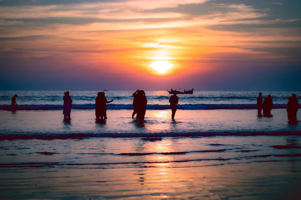 People standing in shallow ocean water at sunset with colorful sky reflections, small boats, and beach silhouettes.