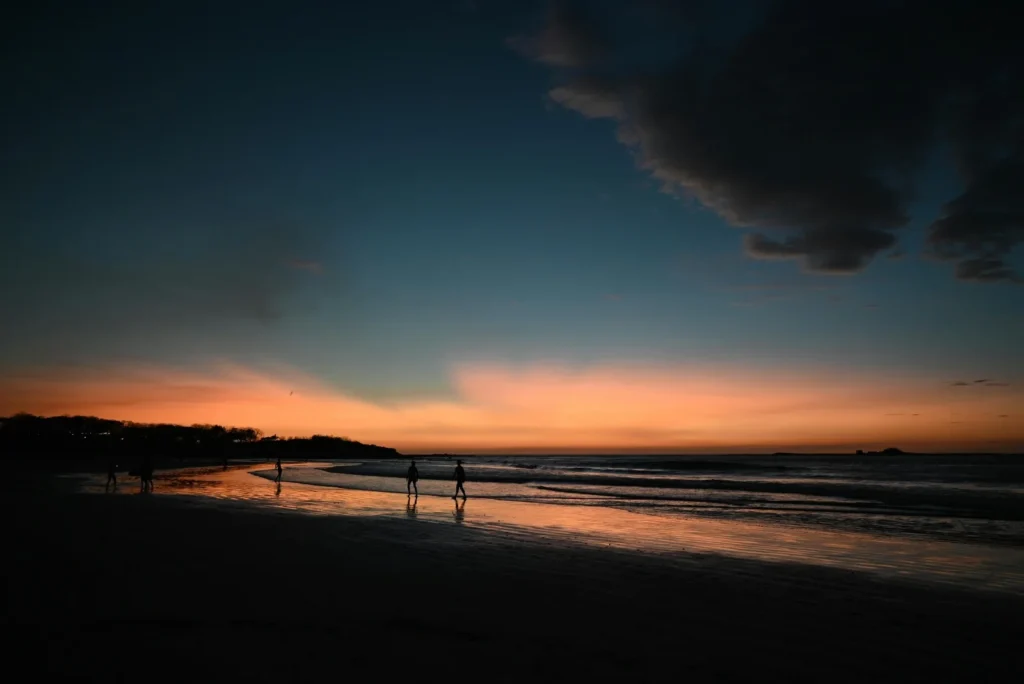People walking along a quiet beach at sunset with orange sky, dark clouds, and waves reflecting on the shore.