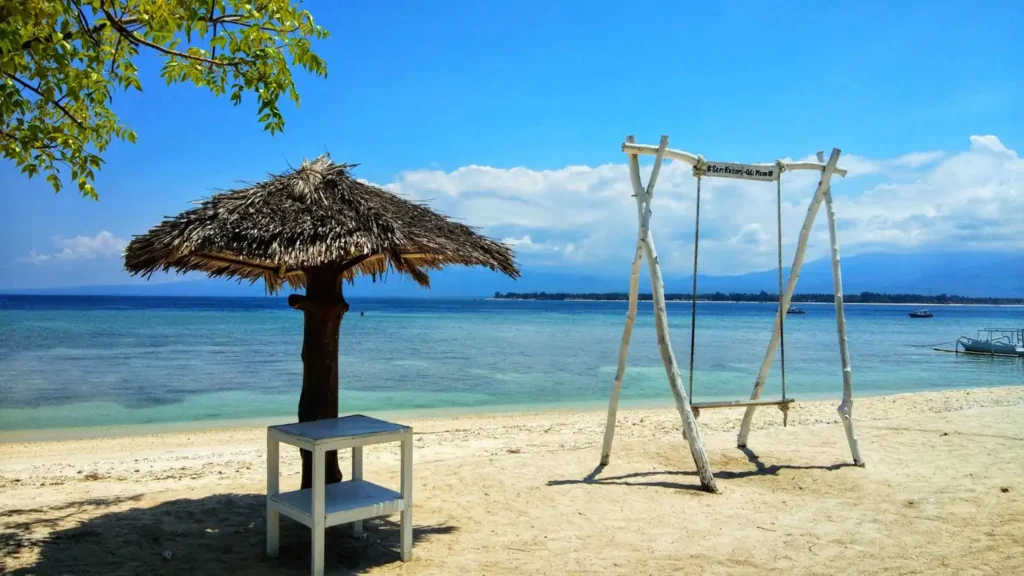 A white wooden beach swing and thatched umbrella on a sandy tropical beach with clear blue water and a bright sky.