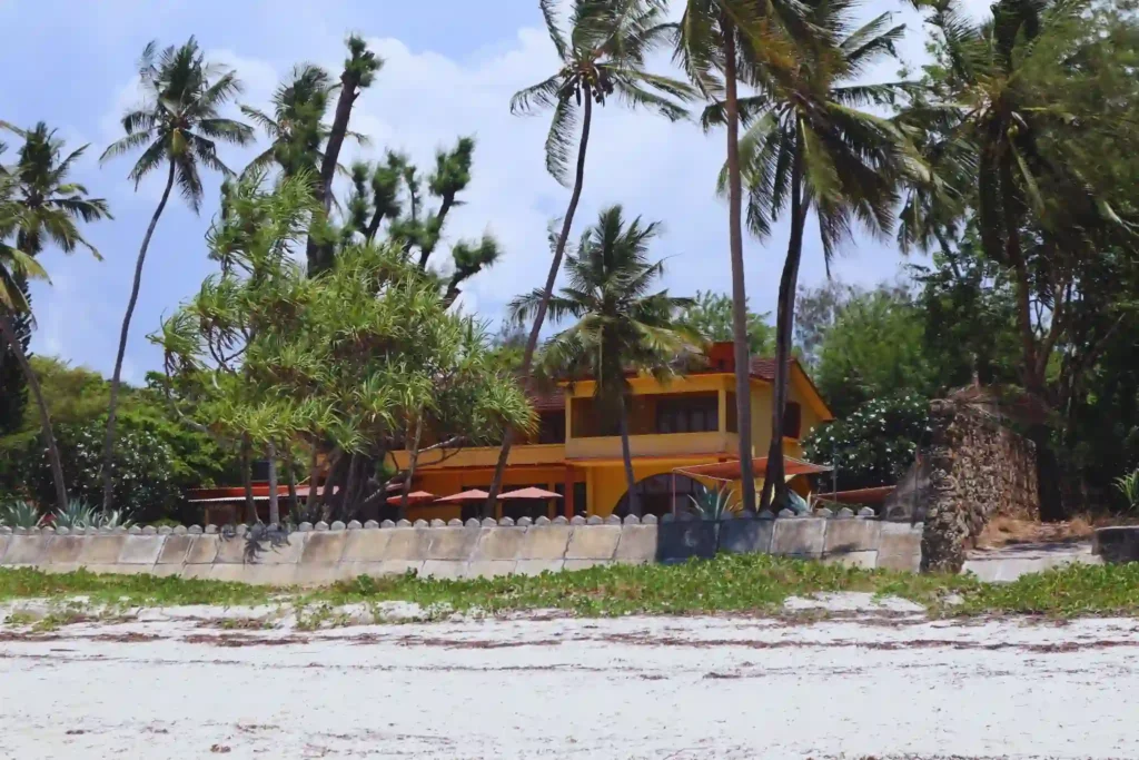 Beachfront tropical villa surrounded by tall palm trees and lush greenery along a white sandy shoreline under a bright sky.