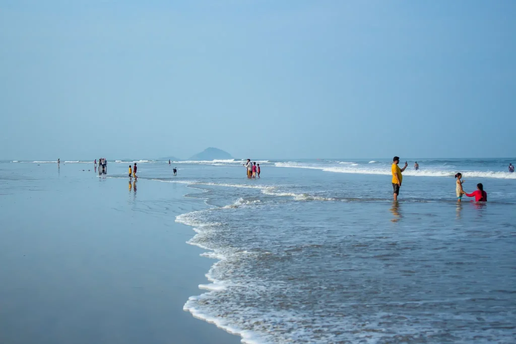 Visitors walking along a wide sandy beach with gentle waves and reflective shoreline under a clear blue sky.