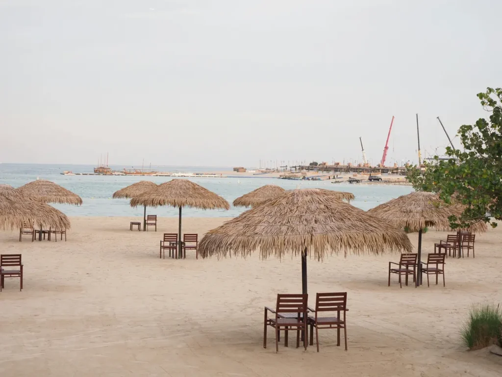 Beach in Qatar with straw umbrellas, wooden chairs, soft sand, and calm sea water near the shoreline.