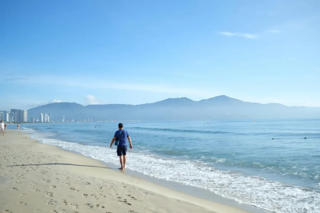 Man walking along a calm sandy beach in Vietnam with blue sea, soft waves, mountains, and city buildings in the distance.