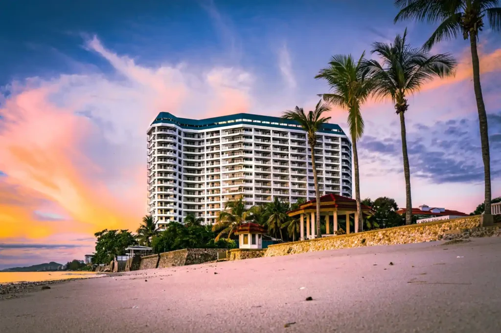 Beachfront hotel with palm trees beside a sandy beach during a colorful sunset sky.
