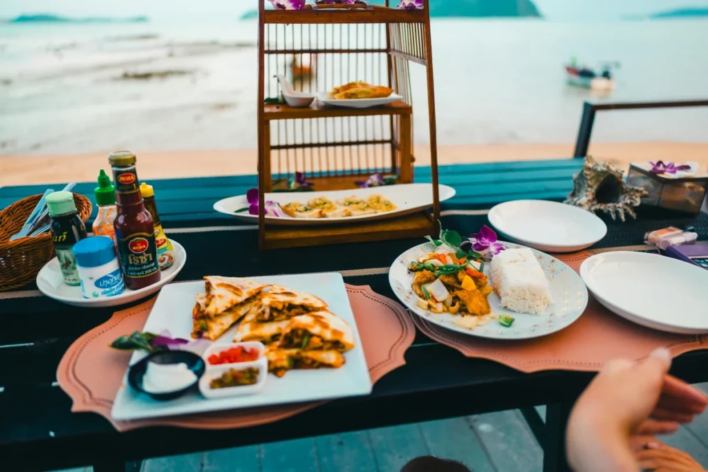Thai food served on a beachfront table with turquoise sea views, plates of rice, snacks, sauces, and tropical flowers